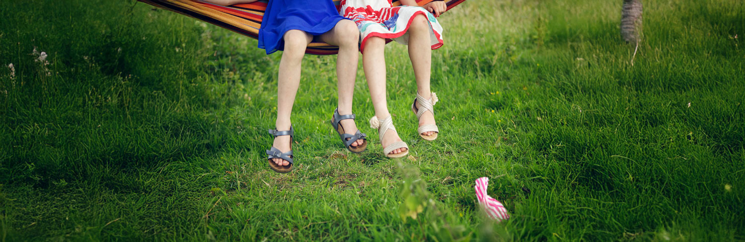 Legs and feet of two girls swinging in a hammock with a bag of sweets on the ground in front of them.